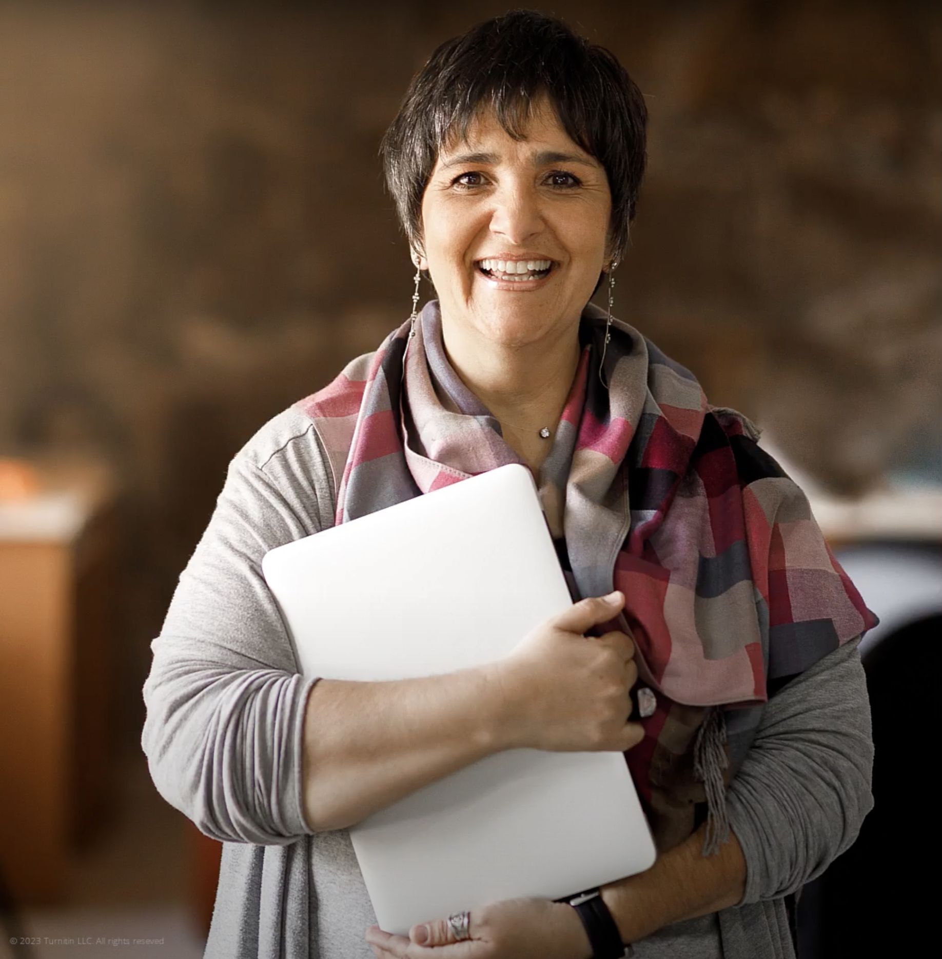 A woman holding a closed laptop against her body smiles into the camera.